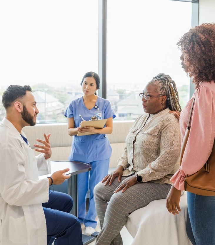 The young adult male doctor gestures as he talks to the mature adult female patient.  The patient's mid adult daughter and the mid adult female nurse pay close attention.